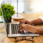 A woman typing on a laptop on a wood table. A green plant is directly behind it, and a table lamp can be seen in the back. There is also a pair of black glasses in the foreground.