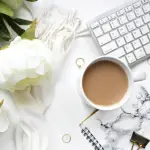 A white table with a white keyboard, a white peony, a marble notebook with a black binder clip on top, and a white coffee cup with coffee inside.