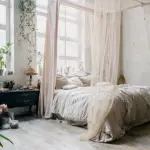 A white bedroom with white canopy drapes, along with green florals in the background.