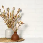 A white vase and a brown vase on a grass matt against a white tile wall. Each vase has light brown and brown wildflowers.