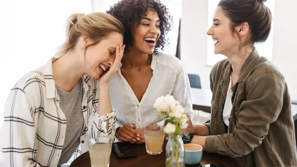 Three women together at a table, having a meaningful conversation. They are drinking coffee and teas, and are laughing together over something that was said by one of the ladies.
